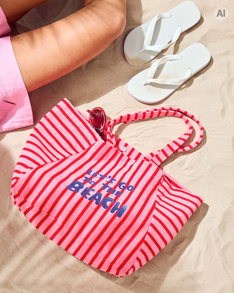 Un sac de plage rayé portant l’inscription « Let’s Go Beach », des lunettes de soleil et des tongs blanches sont disposés sur le sable, créant une ambiance estivale en bord de mer.