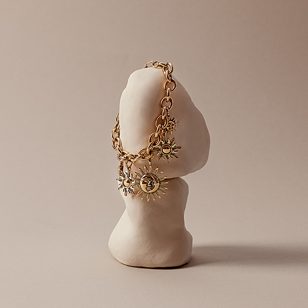 Op een lichte ondergrond ligt een goudkleurige armband met meerdere zonhanger‑bedels, gepresenteerd op een eenvoudige sculptuur.