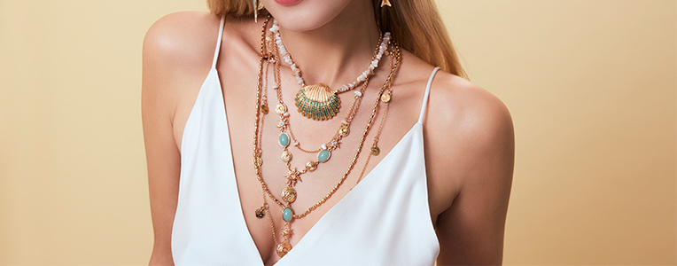 Mehrere goldene Ketten im Retro-Riviera-Stil mit türkisfarbenen Steinen, Perlen und filigranen Anhängern liegen in einem stilvollen Layering-Look auf dem Dekolleté einer Frau.