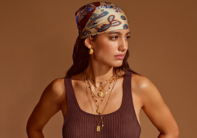 Een persoon staat voor een bruine achtergrond, terwijl de opvallende gouden sieraden en de kleurrijke hoofddoek het geheel een bijzonder karakter geven.