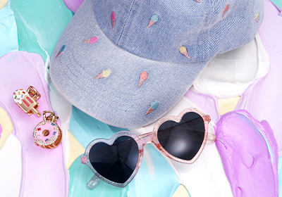 Eine Herzsonnenbrille, zwei bunte Anhänger und eine helle Kappe mit kleinen Eisstickereien liegen auf einem pastellfarbenen Hintergrund.