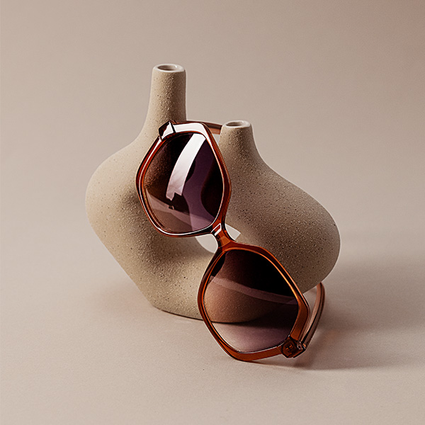 Op een lichte ondergrond leunt een zonnebril met bruine montuur en getinte glazen tegen een beige sculptuur met dubbele vorm.