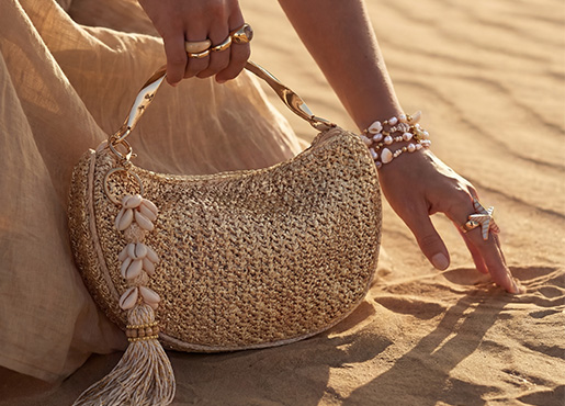  Un bolso se presenta con estilo en la mano de una persona junto con anillos y pulseras sobre un suelo arenoso.