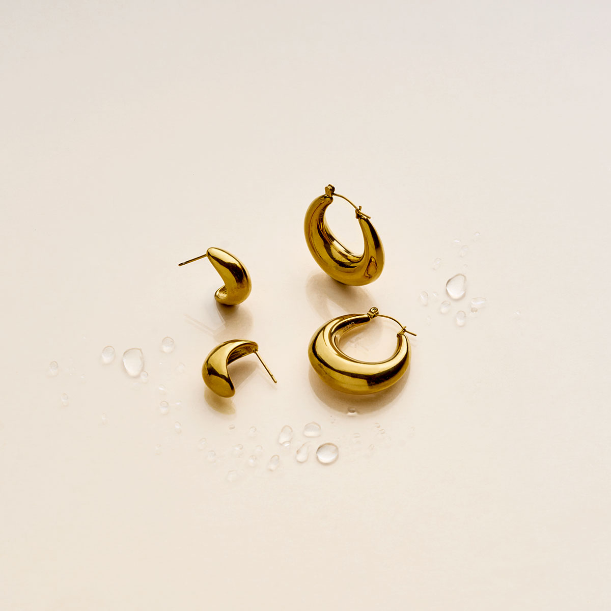 Pendientes de acero inoxidable sobre un fondo beige con gotas de agua.