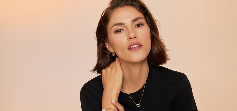 Twee personen dragen fijne gouden sieraden met hartmotief: armbanden en ringen aan de handen, evenals kettingen en oorbellen gecombineerd met spijkerjacks tegen een warme, neutrale achtergrond.