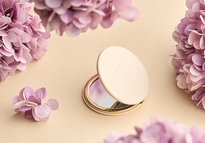 Ein zart schimmernder Kompaktspiegel liegt von sanften, fliederfarbenen Blüten umrahmt und strahlt zeitlose Eleganz sowie feminine Leichtigkeit aus.