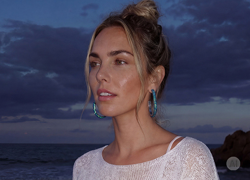 Una persona lleva grandes aros en tonos azul y turquesa y un delicado collar con colgante de concha, combinados con un top blanco de punto frente a un paisaje costero.