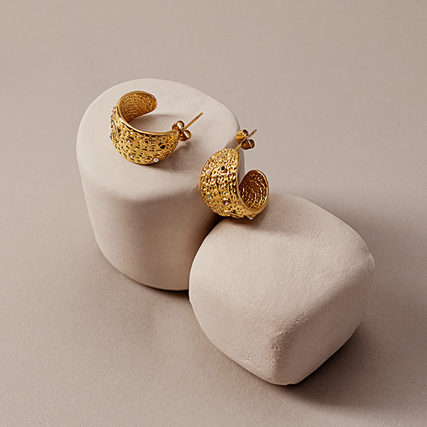 Op een lichte ondergrond staan twee goudkleurige creolen met een gestructureerd oppervlak, gepresenteerd op eenvoudige, afgeronde sokkels.