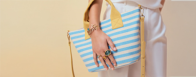 Un bolso a rayas azul y blanco de estilo retro Riviera con detalles dorados se lleva de forma relajada, mientras varios anillos dorados con piedras turquesas destacan la mano de una mujer.