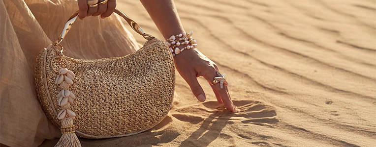  Un bolso se presenta con estilo en la mano de una persona junto con anillos y pulseras sobre un suelo arenoso.