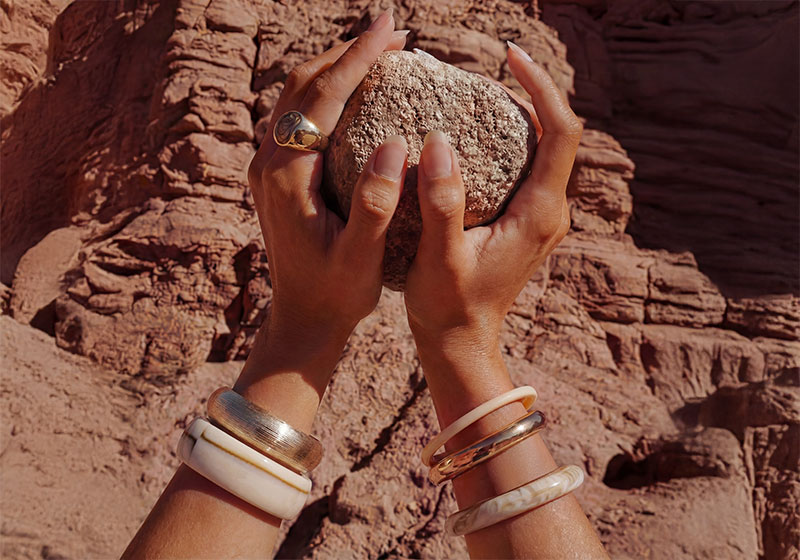  Diversi bracciali rigidi sono presentati con stile su mani che tengono una pietra davanti a un caldo paesaggio desertico roccioso.