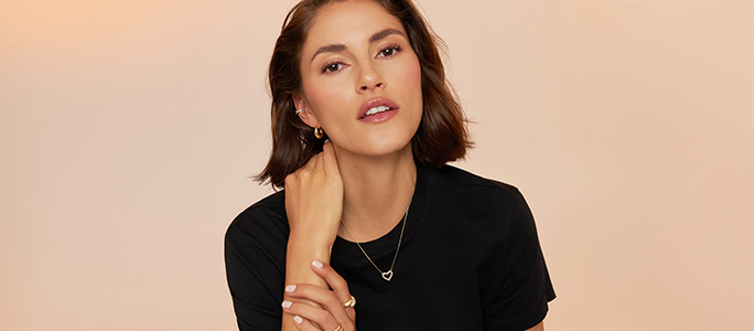 Twee personen dragen fijne gouden sieraden met hartmotief: armbanden en ringen aan de handen, evenals kettingen en oorbellen gecombineerd met spijkerjacks tegen een warme, neutrale achtergrond.